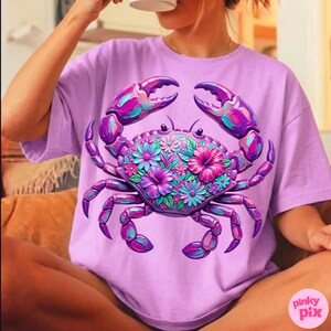 May include: A lilac-colored t-shirt featuring a vibrant crab design. The crab is adorned with colorful flowers in shades of pink, purple, and teal. The crab's claws and legs are a deep purple, creating a striking contrast. The shirt has a relaxed fit.