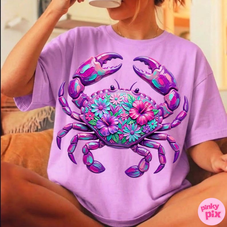 May include: A lilac-colored t-shirt featuring a vibrant crab design. The crab is adorned with colorful flowers in shades of pink, purple, and teal. The crab's claws and legs are a deep purple, creating a striking contrast. The shirt has a relaxed fit.