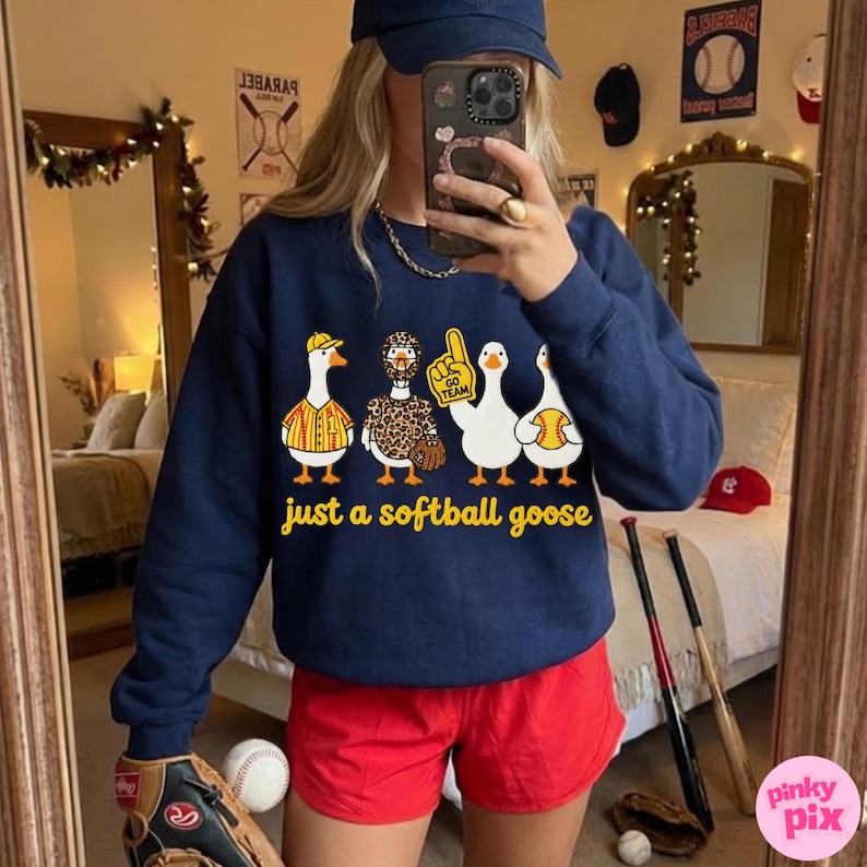 May include: Navy blue sweatshirt with a softball theme. The graphic features four cartoon geese in baseball-themed outfits, with the text "just a softball goose" below. The person is wearing red shorts and a baseball cap.