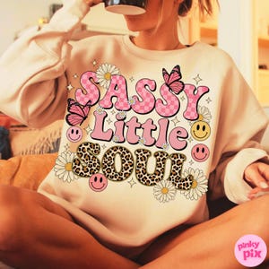 May include: A beige sweatshirt with the words "SASSY Little SOUL" in pink, checkered, and leopard print lettering. The design includes pink butterflies, daisies, and smiley faces. The sweatshirt is a casual, comfortable style.