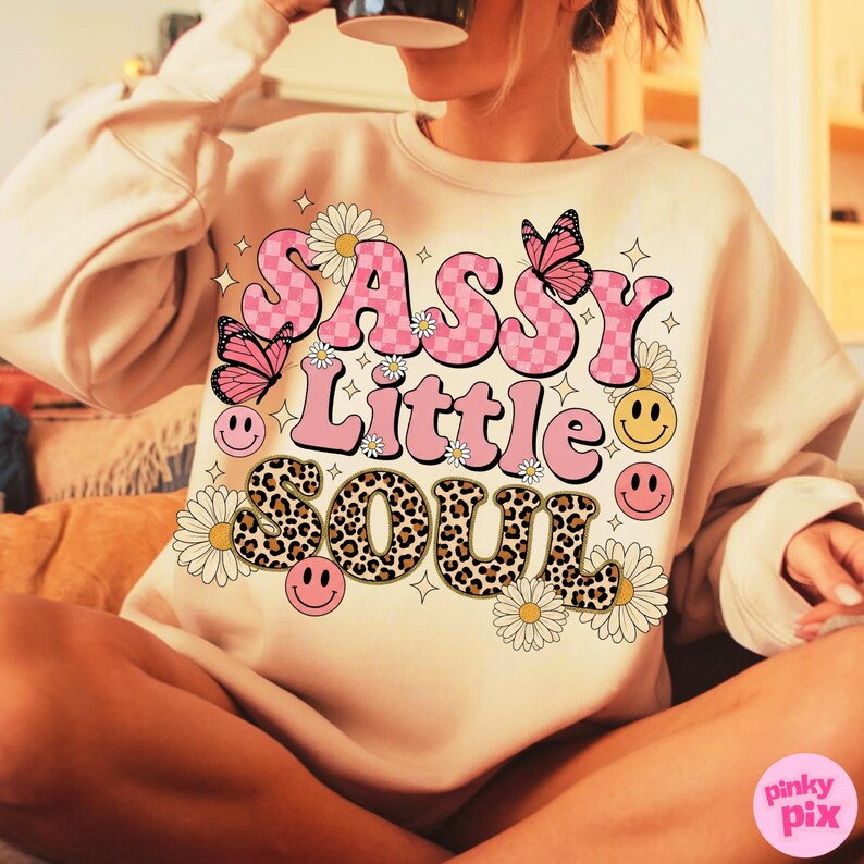 May include: A beige sweatshirt with the words "SASSY Little SOUL" in pink, checkered, and leopard print lettering. The design includes pink butterflies, daisies, and smiley faces. The sweatshirt is a casual, comfortable style.