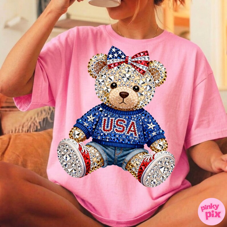 May include: Pink t-shirt featuring a bejeweled teddy bear design. The bear wears a blue sweater with "USA" in red letters, denim shorts, and red, white, and blue sneakers. A matching bow adorns the bear's head. The design is embellished with sparkling crystals.