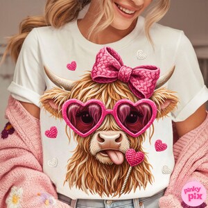 May include: White t-shirt featuring a whimsical illustration of a Highland cow wearing pink heart-shaped sunglasses and a matching bow. The cow has its tongue out and is holding a heart-shaped lollipop. Scattered pink and white hearts complete the design.