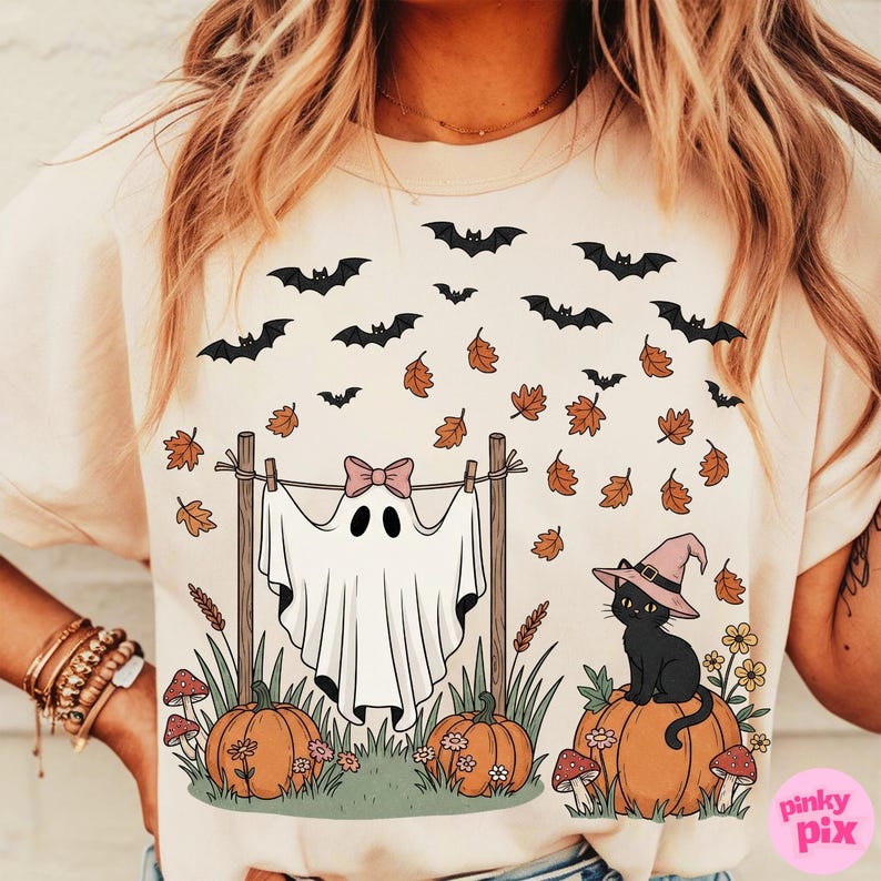 May include: Beige t-shirt with a Halloween-themed graphic. The design features a ghost hanging on a clothesline with a pink bow, pumpkins, a black cat wearing a witch hat, bats, and autumn leaves. The shirt has a round neckline and short sleeves.