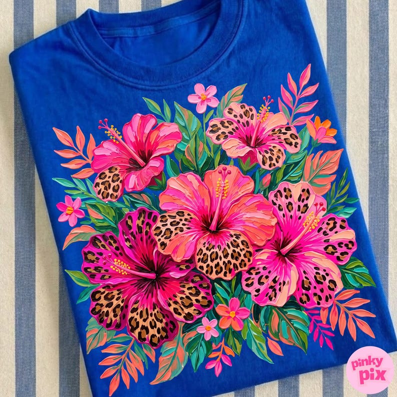 May include: A royal blue t-shirt featuring a vibrant floral design. The design includes large pink hibiscus flowers with leopard print centers, surrounded by green leaves and smaller pink flowers. The shirt has a crew neck.