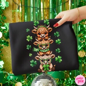 May include: Black tote bag featuring a stack of three cartoon Highland cows, each adorned with shamrocks and green accessories. The cows wear glasses and scarves, with shamrocks scattered around. The design is festive and themed for St. Patrick's Day.