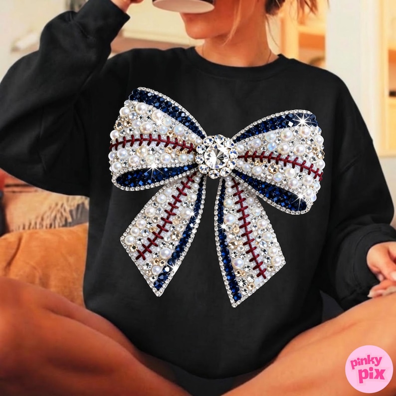 May include: Black sweatshirt featuring a large bow design. The bow is embellished with white pearls, blue and red accents, and a central diamond-like jewel. The bow's design resembles a baseball stitch pattern. The sweatshirt is a casual apparel item.