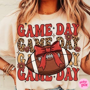 May include: Cream-colored t-shirt with a football graphic and the words "GAME DAY" in a retro font. The football has a red bow and the design includes floral accents and leopard print.