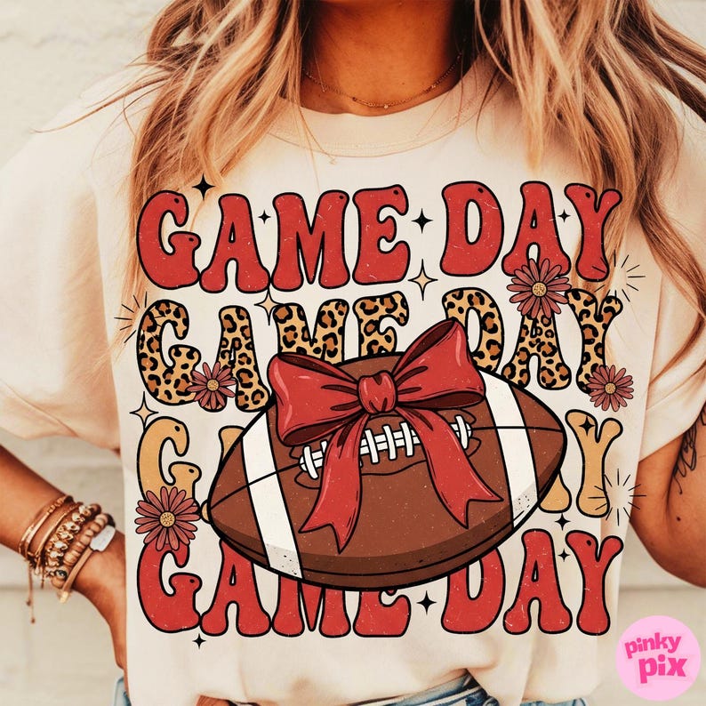 May include: Cream-colored t-shirt with a football graphic and the words "GAME DAY" in a retro font. The football has a red bow and the design includes floral accents and leopard print.