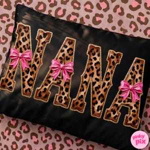 May include: Black pillow with the word "NANA" in leopard print letters outlined in gold glitter. Each letter has a pink bow. The pillow is on a pink and brown leopard print background.