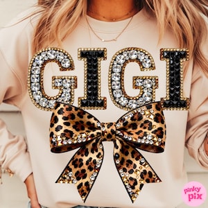 May include: A light beige sweatshirt with the word "GIGI" in large, jeweled letters. The letters are outlined in gold and filled with clear rhinestones and black gems. A leopard print bow with rhinestones is below the text.