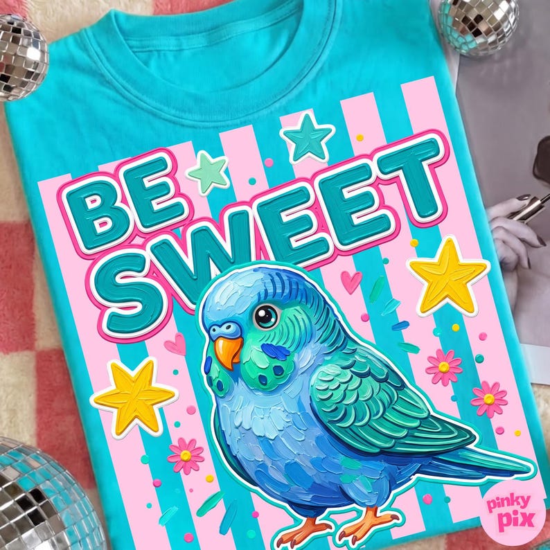 May include: A turquoise t-shirt with a graphic that says "BE SWEET" in a bubble font. The graphic includes a blue parakeet, pink stripes, stars, and flowers. The shirt has a round neckline.