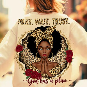 Pu&ograve; includere: Felpa color crema con grafica di una donna con foulard e abiti leopardati, circondata da rose rosse e glitter dorati. Il testo "PRAY. WAIT. TRUST." &egrave; in alto e "God has a plan" in basso.