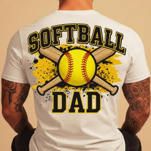 May include: White t-shirt with a softball-themed graphic. The design features a yellow softball, crossed baseball bats, and the words "SOFTBALL DAD" in a bold, black and yellow font. The design is on the back of the shirt.
