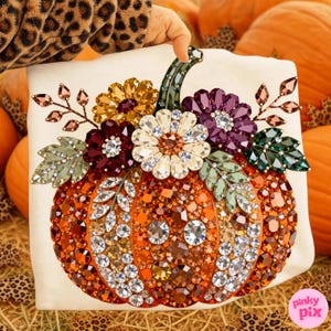 May include: A white sweatshirt featuring a large pumpkin graphic embellished with faceted crystals in shades of orange, brown, and clear. The pumpkin is topped with a cluster of crystal flowers in yellow, purple, and white, accented by green crystal leaves.