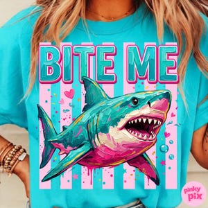 May include: A turquoise t-shirt with the words "BITE ME" in large, teal letters. A colorful shark illustration with pink and teal accents is centered below the text. Pink and white stripes and hearts complete the design.