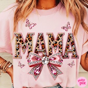 May include: Pink t-shirt with the word "MAMA" in leopard print letters, outlined with rhinestones. A large pink bow with rhinestones is below the word, and pink butterfly accents are scattered around the design. The shirt is a casual style.