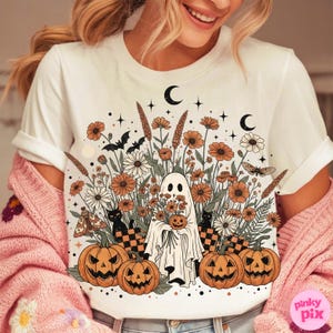 May include: White t-shirt with a Halloween design. The graphic shows a ghost with a pumpkin, flowers, pumpkins, bats, and moons. The colors are orange, black, and white. The design has a vintage, autumnal feel.