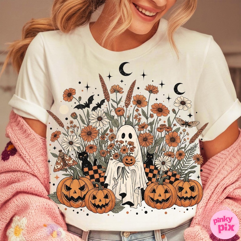 May include: White t-shirt with a Halloween design. The graphic shows a ghost with a pumpkin, flowers, pumpkins, bats, and moons. The colors are orange, black, and white. The design has a vintage, autumnal feel.