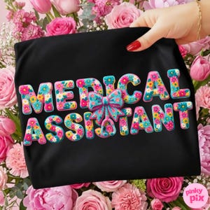 May include: Black t-shirt with the words "MEDICAL ASSISTANT" in colorful floral letters. A blue and pink bow and stethoscope graphic is centered between the words. The shirt is held up by a hand with red nail polish, surrounded by pink flowers.