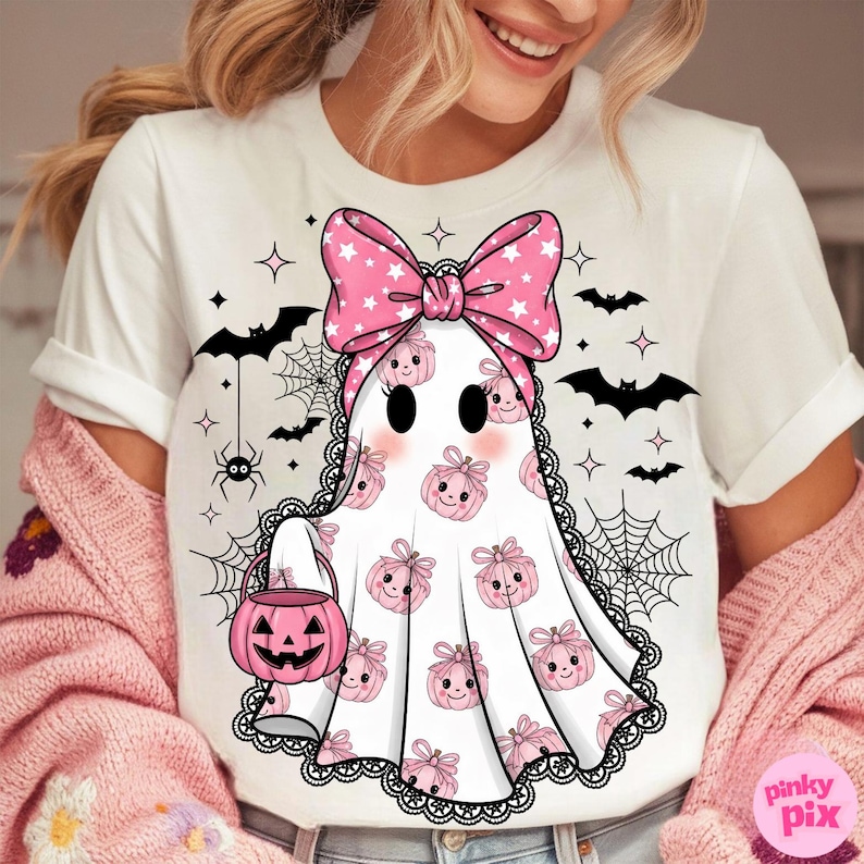 May include: White t-shirt with a ghost design, a pink bow, and pumpkin details. The design includes bats, spiderwebs, and stars, ideal for Halloween. The ghost holds a pink jack-o'-lantern bucket.