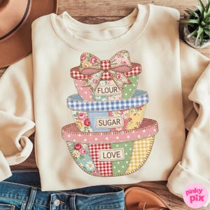 May include: Cream-colored sweatshirt featuring a stack of patchwork bowls with the words "FLOUR", "SUGAR", and "LOVE". The bowls have floral, gingham, and polka dot patterns. A large bow sits atop the top bowl.