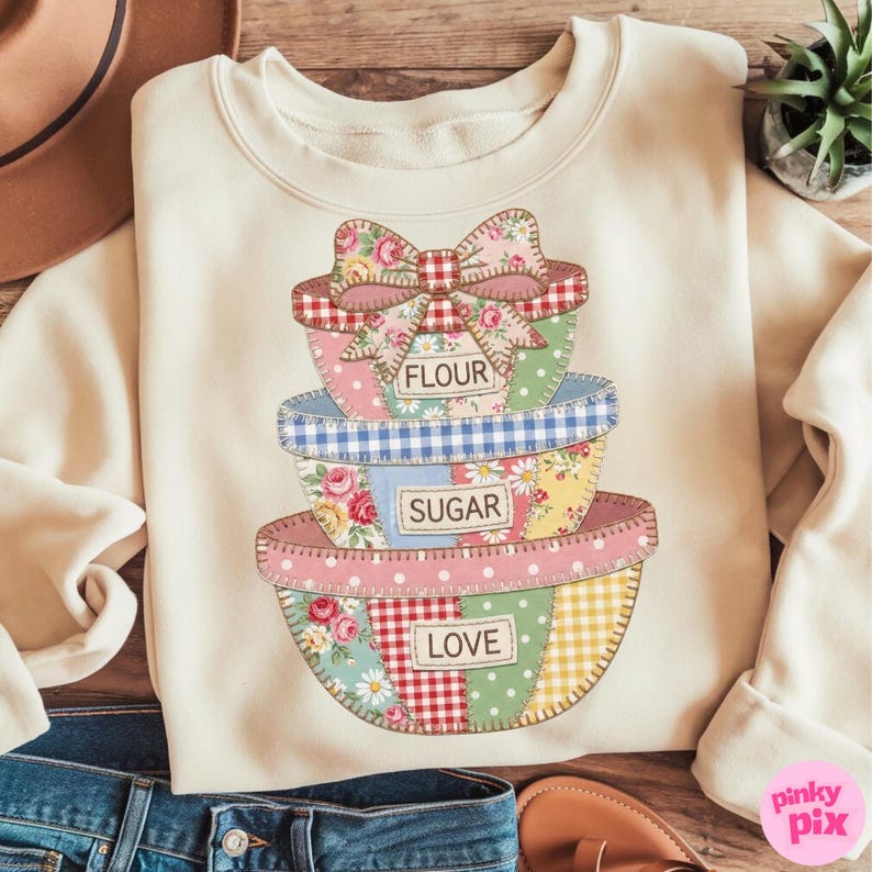 May include: Cream-colored sweatshirt featuring a stack of patchwork bowls with the words "FLOUR", "SUGAR", and "LOVE". The bowls have floral, gingham, and polka dot patterns. A large bow sits atop the top bowl.