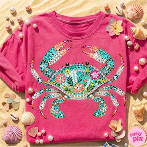 May include: A hot pink t-shirt featuring a detailed crab design. The crab is adorned with turquoise, white, and pink gems, and floral accents. The design is set against a sandy background with seashells and pearls.
