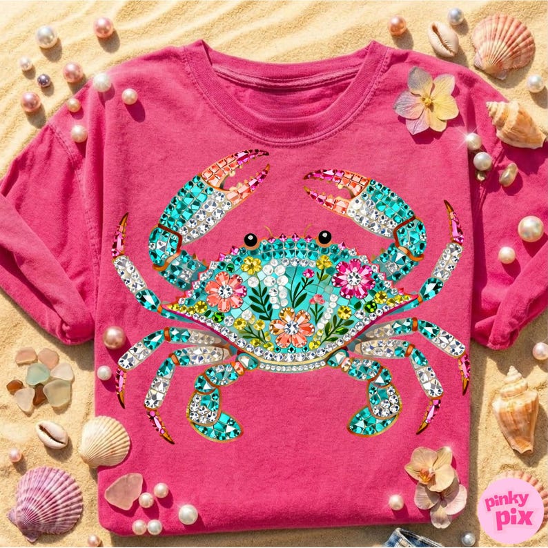 May include: A hot pink t-shirt featuring a detailed crab design. The crab is adorned with turquoise, white, and pink gems, and floral accents. The design is set against a sandy background with seashells and pearls.