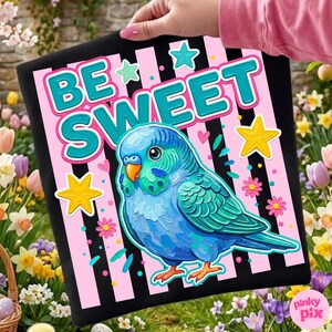 May include: A black square with the words "BE SWEET" in teal and white, and a blue parakeet illustration. The background has pink stripes and star and flower accents. The image is held in front of a garden.