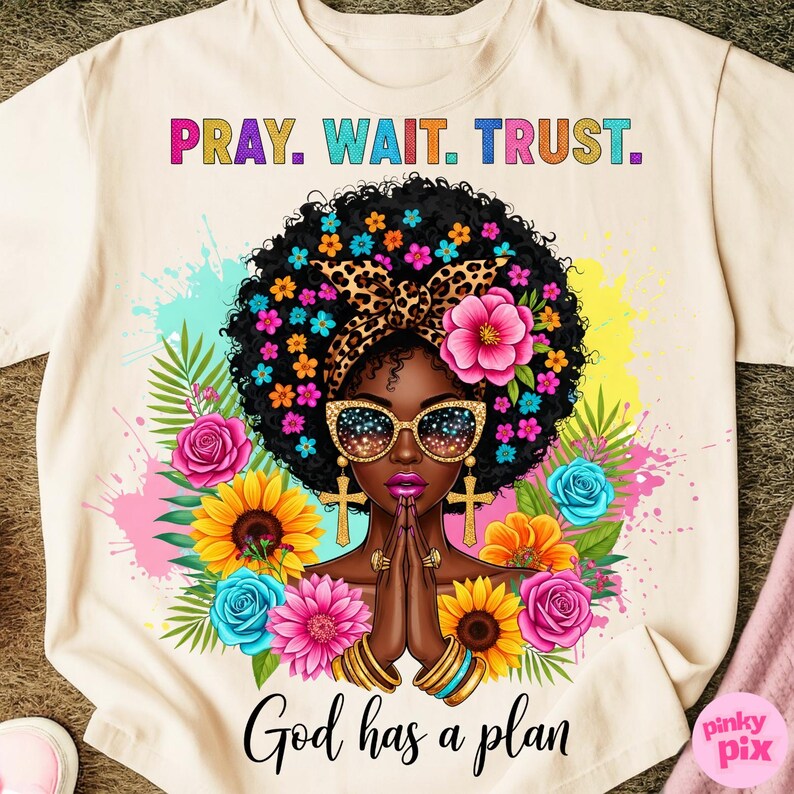 May include: Cream-colored t-shirt featuring a vibrant graphic of a Black woman with sunglasses, floral accents, and the text "PRAY. WAIT. TRUST." and "God has a plan." The design includes colorful flowers and a leopard print headband.