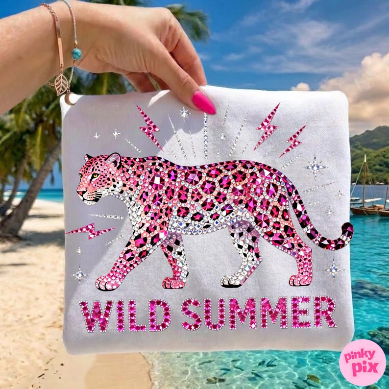 May include: White t-shirt featuring a pink leopard design with rhinestone accents. The leopard is walking, surrounded by lightning bolts and stars. The words "WILD SUMMER" are printed in pink. The background shows a beach scene.