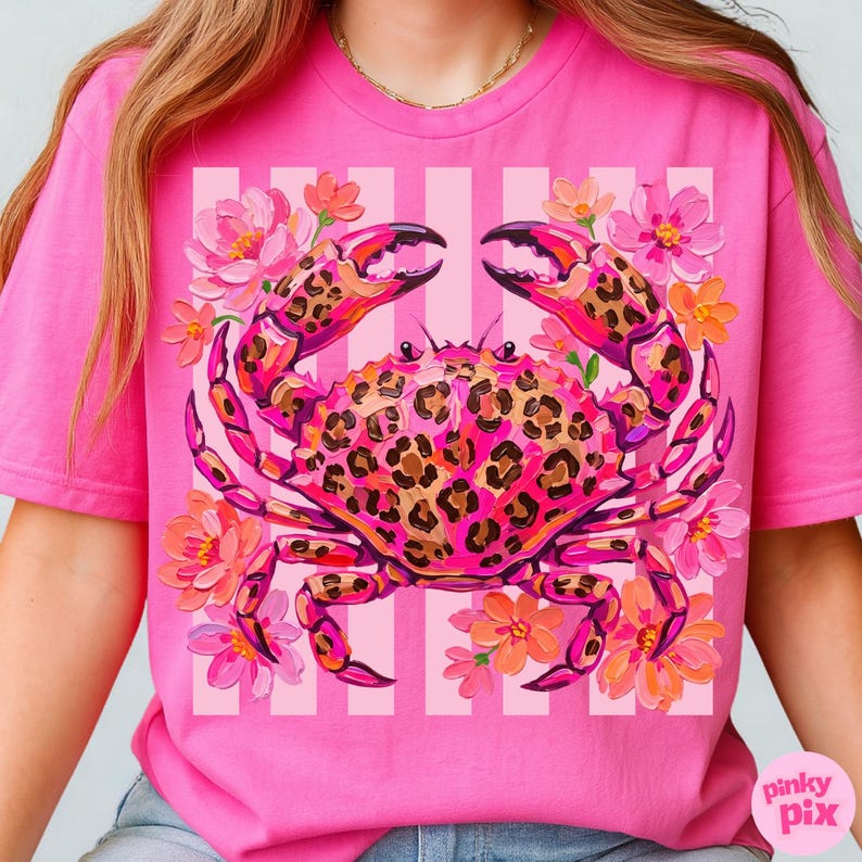 May include: Pink t-shirt featuring a crab design with leopard print details. The crab is surrounded by pink and orange flowers, set against a striped background. The shirt has a crew neck and short sleeves.