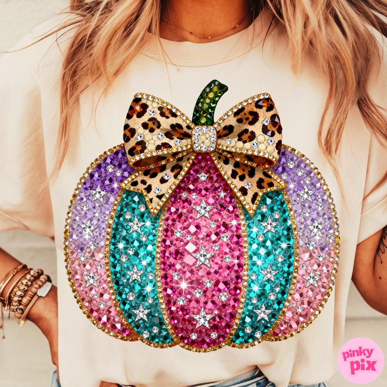 Op de afbeelding: Een cr&egrave;mekleurig t-shirt met een grote afbeelding van een pompoen versierd met roze, turquoise en paarse strass-steentjes, geaccentueerd door zilveren stervormige studs en een strik met luipaardprint met een groene strass-steel.