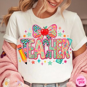May include: White t-shirt with the word "TEACHER" in colorful, whimsical lettering. The design includes a bow with an apple, a pencil, stars, and floral accents. The shirt is a casual, everyday wear item.