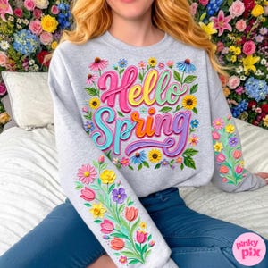 May include: A light grey sweatshirt featuring the words "Hello Spring" in colourful, 3D lettering, surrounded by floral designs. The sleeves also have floral patterns. The sweatshirt is worn with blue jeans.