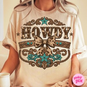 Pu&ograve; includere: Una maglietta beige con un design a stampa di mucca marrone e bianca che dice "HOWDY" in stile vintage con un fiocco turchese e stelle.