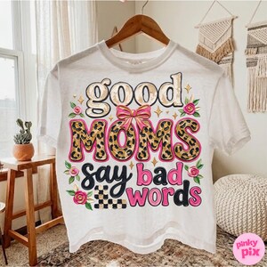 May include: White t-shirt with the phrase "good moms say bad words" in a playful design. The word "moms" is in a leopard print pattern, and the shirt is decorated with pink roses and a checkered pattern.