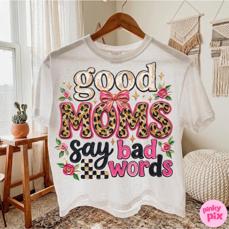 May include: White t-shirt with the phrase "good moms say bad words" in a playful design. The word "moms" is in a leopard print pattern, and the shirt is decorated with pink roses and a checkered pattern.