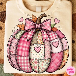 May include: Cream-colored sweatshirt featuring a patchwork pumpkin design. The pumpkin is composed of various pink patterns, including gingham, floral, and polka dots, with a large pink bow and heart accents. The design has a fall theme.