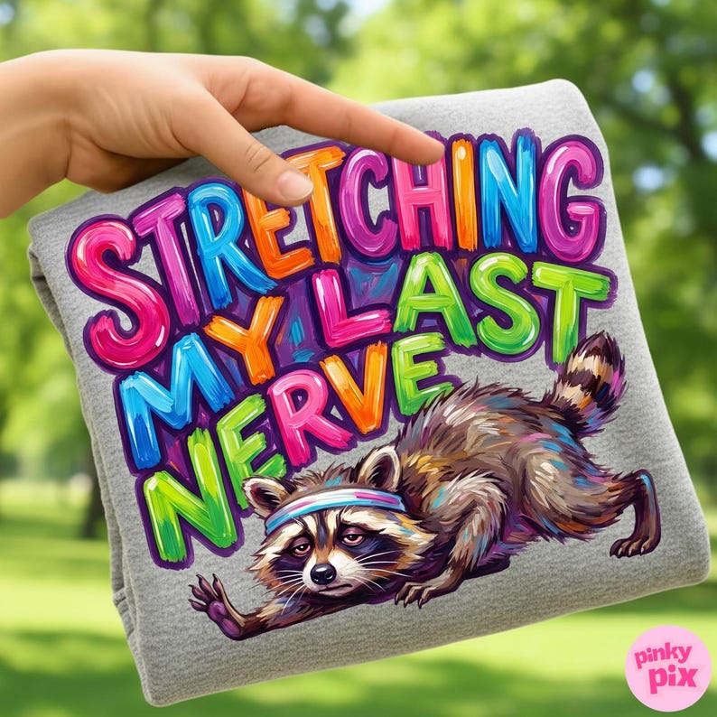 May include: Gray t-shirt with the phrase "STRETCHING MY LAST NERVE" in colorful bubble letters. A cartoon raccoon wearing a headband is stretching. The t-shirt is held up by a hand against a blurred green background.