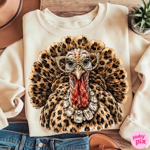 May include: A cream-colored sweatshirt featuring a graphic print of a turkey with leopard print feathers. The turkey wears leopard print glasses and a large, clear crystal-style statement necklace. The design is centered on the front of the garment.