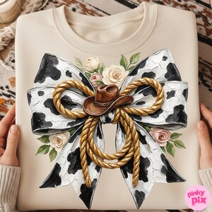 May include: Cream-colored sweatshirt featuring a large bow design with a cow print pattern in black and white. A brown cowboy hat sits in the center, surrounded by a gold rope and floral accents. The design is detailed and artistic.