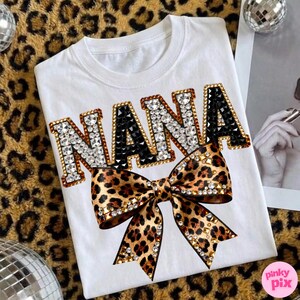 May include: White t-shirt with the word "NANA" in jeweled letters, black and clear rhinestones, and a leopard print bow. The shirt has a crew neck and is likely made of cotton or a similar fabric.