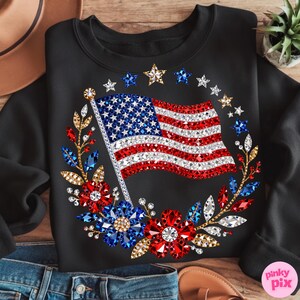 May include: Black sweatshirt featuring a sparkling American flag design. The flag is embellished with red, white, and blue crystals, surrounded by a wreath of flowers and stars. A patriotic themed apparel item.