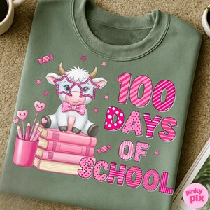 Pu&ograve; includere: Felpa verde salvia con una mucca dei cartoni animati con occhiali a forma di stella e un papillon, seduta su libri rosa. Il testo "100 GIORNI DI SCUOLA" &egrave; in rosa. Matite rosa e lecca-lecca a forma di cuore completano il design.
