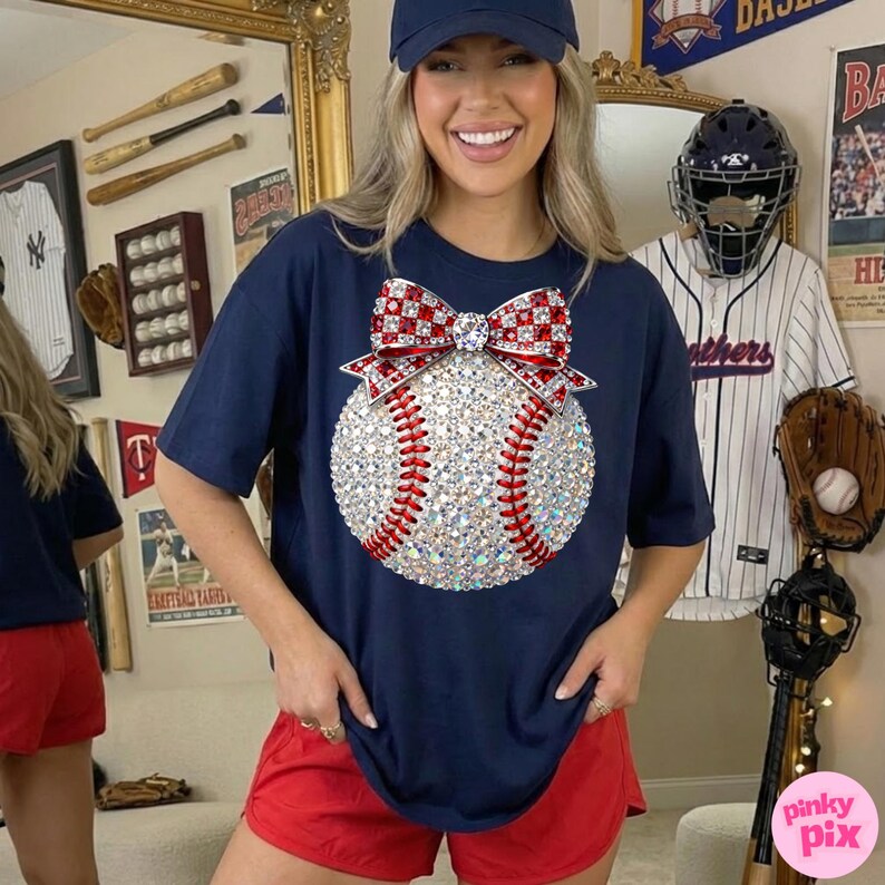 May include: Navy blue t-shirt featuring a baseball design. The baseball is embellished with sparkling rhinestones and red stitching, topped with a red and white bow. The person is wearing red shorts and a navy blue baseball cap.