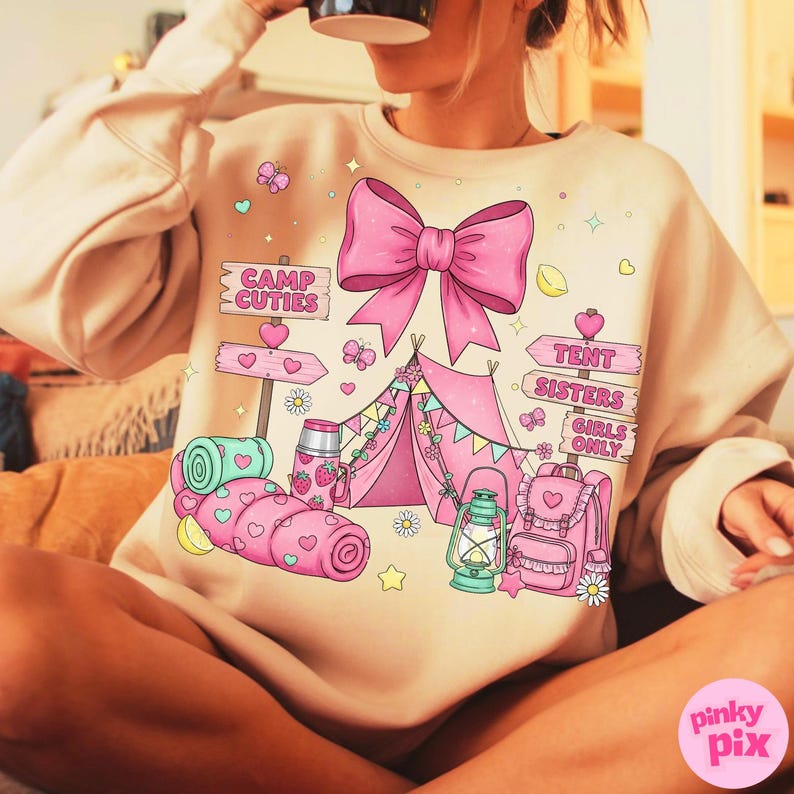 May include: A beige sweatshirt featuring a pink camping design. The graphic includes a pink tent, bow, backpack, sleeping bag, and signs that read "Camp Cuties" and "Tent Sisters Girls Only."