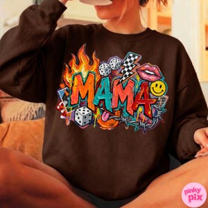 May include: A brown sweatshirt with a colorful graphic design that says "MAMA". The design includes flames, dice, a lightning bolt, lips, a smiley face, and barbed wire. The word "MAMA" is in a graffiti style font.