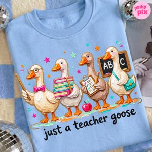 May include: Light blue sweatshirt featuring a whimsical illustration of four geese dressed as teachers. One goose holds a pointer, another carries books, a third has a clipboard, and the last holds a chalkboard. The text "just a teacher goose" is below.